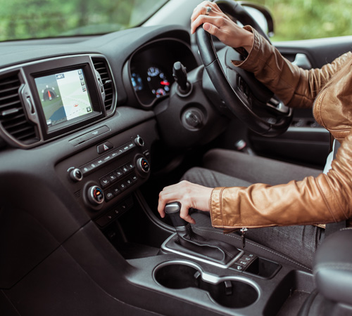 Close-up, woman driving a car includes a gearbox, reversing, starting to move in woods, leather jacket, automatic transmission. Touch screen navigation map app on monitor.